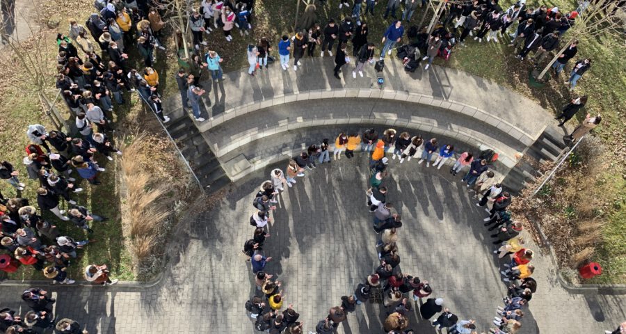 Schulangehörige befinden sich auf dem Schulhof und bilden vor einem Treppenaufgang zusammen ein Peace Zeichen