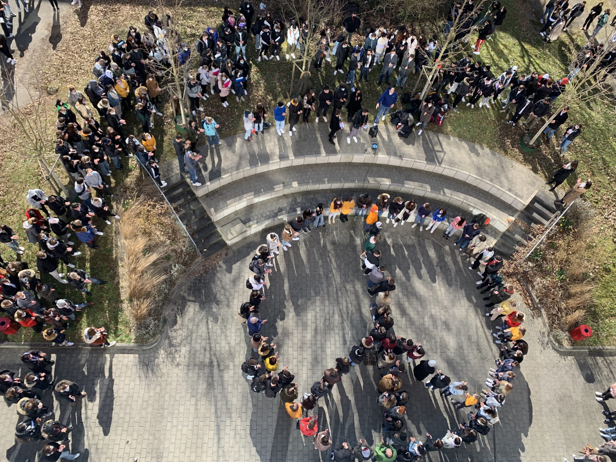 Schulangehörige befinden sich auf dem Schulhof und bilden vor einem Treppenaufgang zusammen ein Peace Zeichen